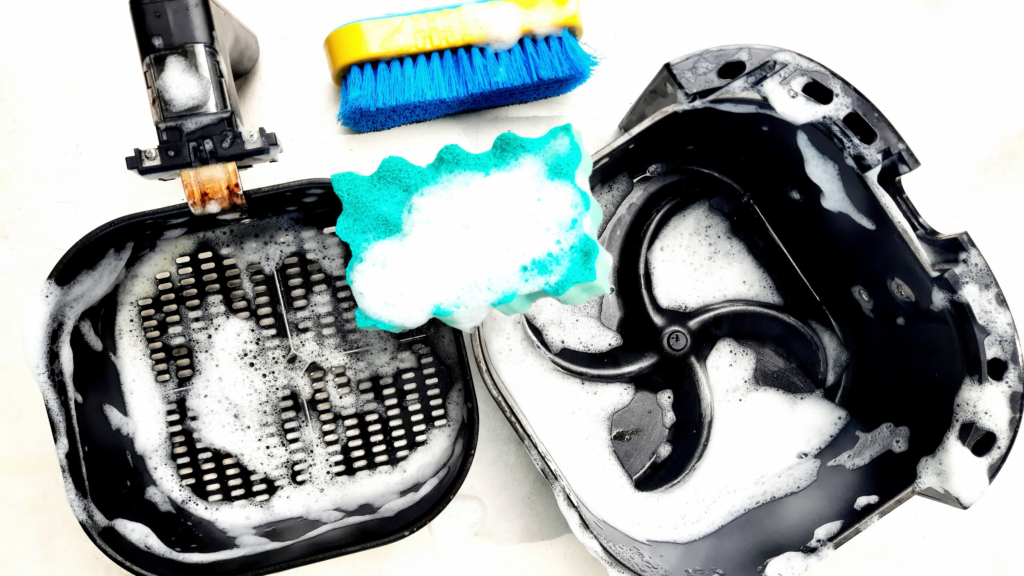 a tray and a basket of an air fryer covered in soap, a blue sponge with the soap foam on it